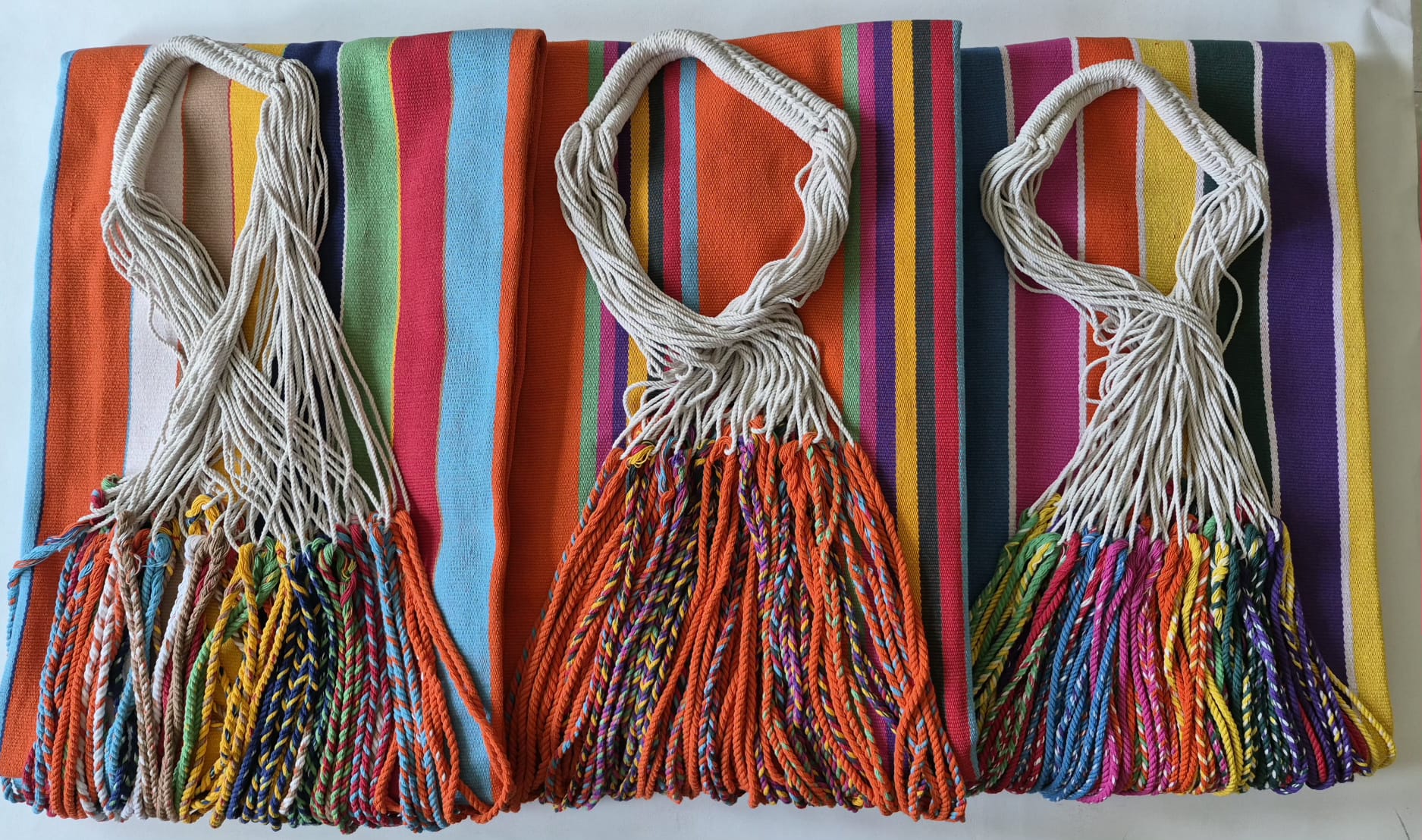 Three folded handwoven hammocks with colorful stripes, rope loops and braided fringes arranged side by side
