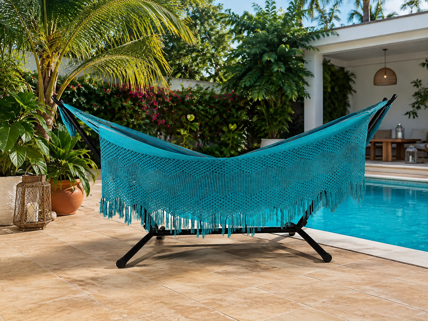 Turquoise handwoven hammock with fringe on a black stand beside a swimming pool in a tropical garden
